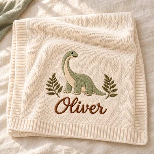 Digital design preview: cream knit blanket with a long-neck dinosaur, two leafy branches and the example embroidered name Oliver.